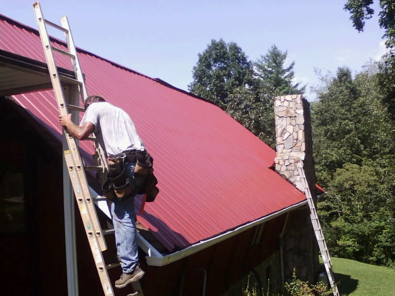 Roofer on a ladder beside a red metal roof and stone chimney — Soffit & Fascia Repair by Roofing Brothers in Hill City